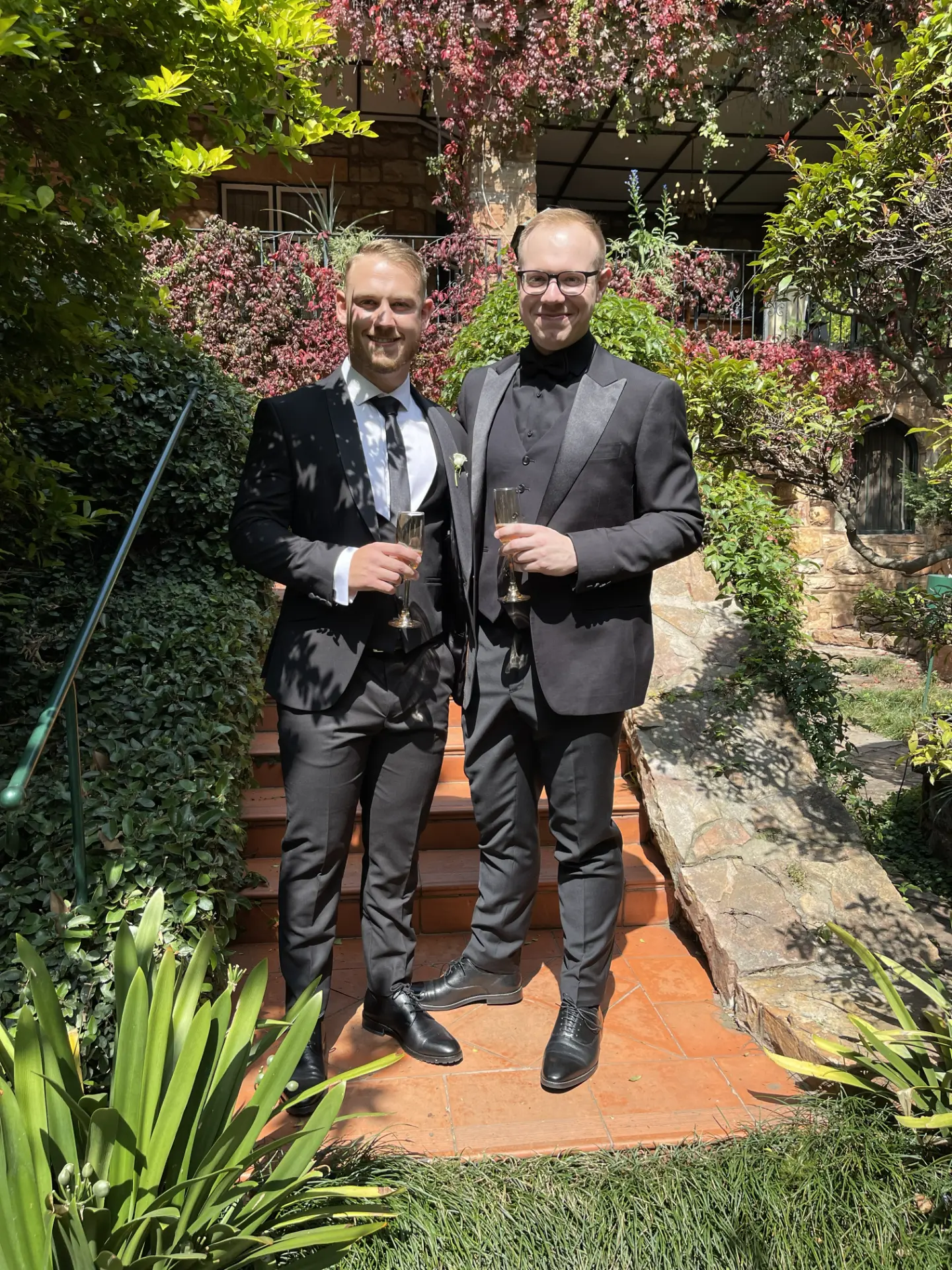 Jason Knowles and Devan Sequeira in suits holding champagne glasses in a garden